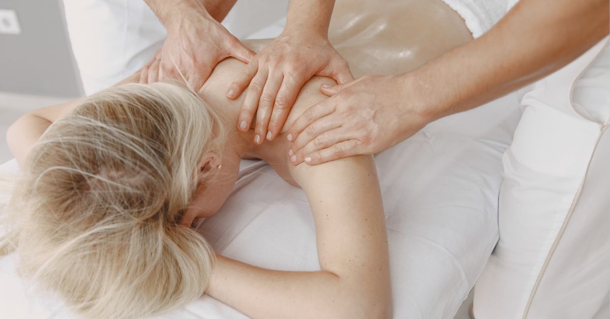A Blonde Woman Lying Face Down On A Professional Massage Table Covered With A White Towel While Receiving A Back Massage From A Therapist